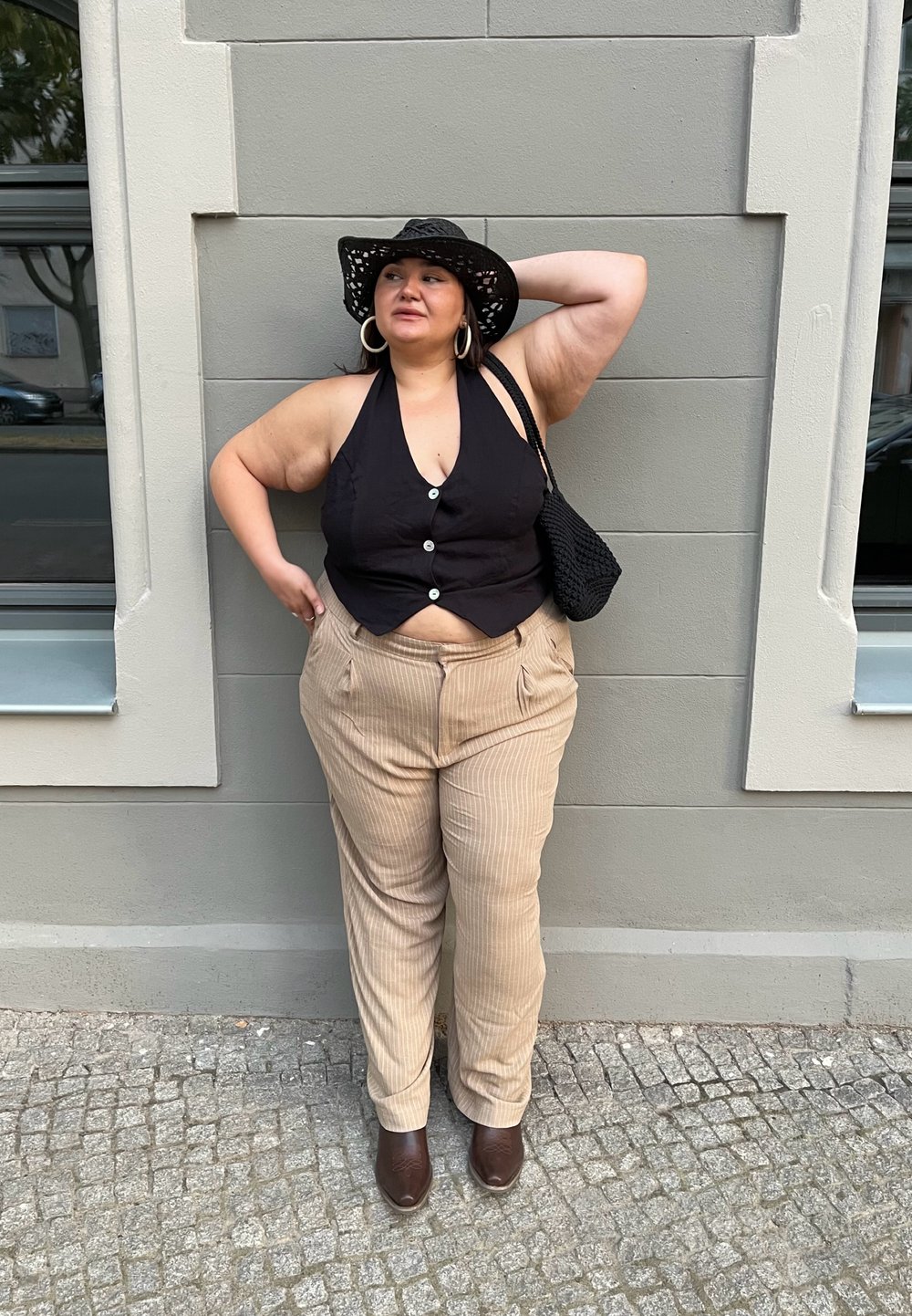 Schwarzes ärmelloses Oberteil mit Knöpfen, beigefarbene Nadelstreifenhose, braune Stiefeletten und einen schwarzen geflochtenen Hut. Stehend vor einer grauen Wand.