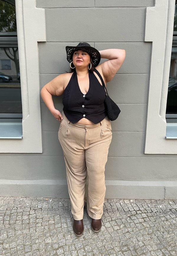 Top negro sin mangas con botones, pantalones beige a rayas, botas marrones hasta el tobillo y un sombrero negro tejido. De pie contra una pared gris.