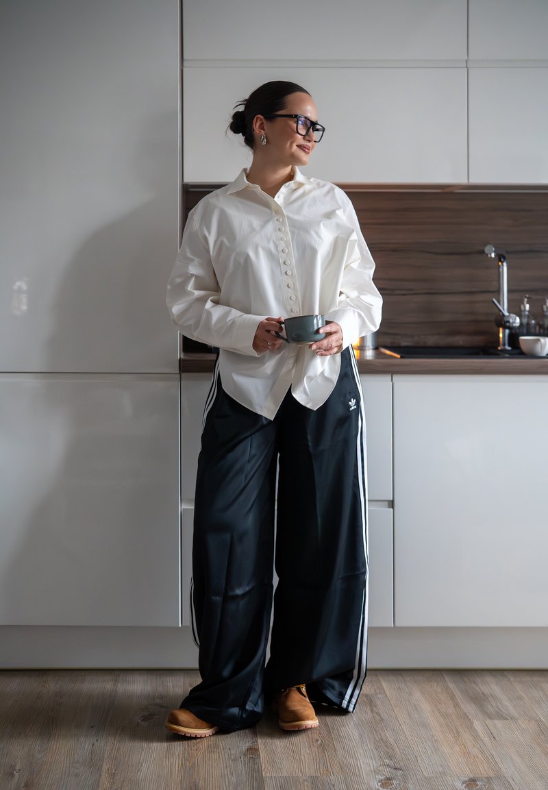 Vrouw met een bril en casual kleding staat in een moderne keuken, houdt een mok vast, kijkt opzij en glimlacht.