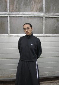Schwarzer Sweatshirt mit Reißverschluss und weißem Logo, kombiniert mit lockeren schwarzen Hosen mit weißen Seitenstreifen. Haare zurückgebunden, steht vor einer strukturierten Wand.