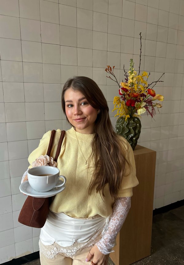 Jeune femme en pull jaune tenant une tasse de thé et une soucoupe, debout près d'une table en bois avec un vase de fleurs colorées.