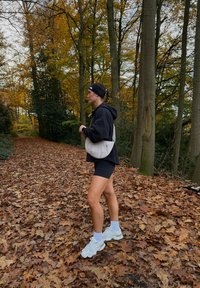 Treningsklær inkluderer en svart genser og shorts. En lysfarget skulderveske bæres over skulderen. Bakgrunnen har høstløvverk.