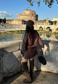 Bruin sportief outfit met blauwe strepen; hoge kniehoge bruine laarzen; hoofddoek met luipaardprint; staand bij een stenen balustrade met een kasteel op de achtergrond.
