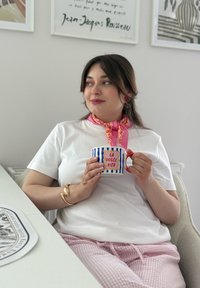 Tazza con strisce blu e bianche, manico rosso e testo "LA DOLCE VITA". Persona indossa una camicia bianca e pantaloni da pigiama a fantasia rosa. Sfondo chiaro.