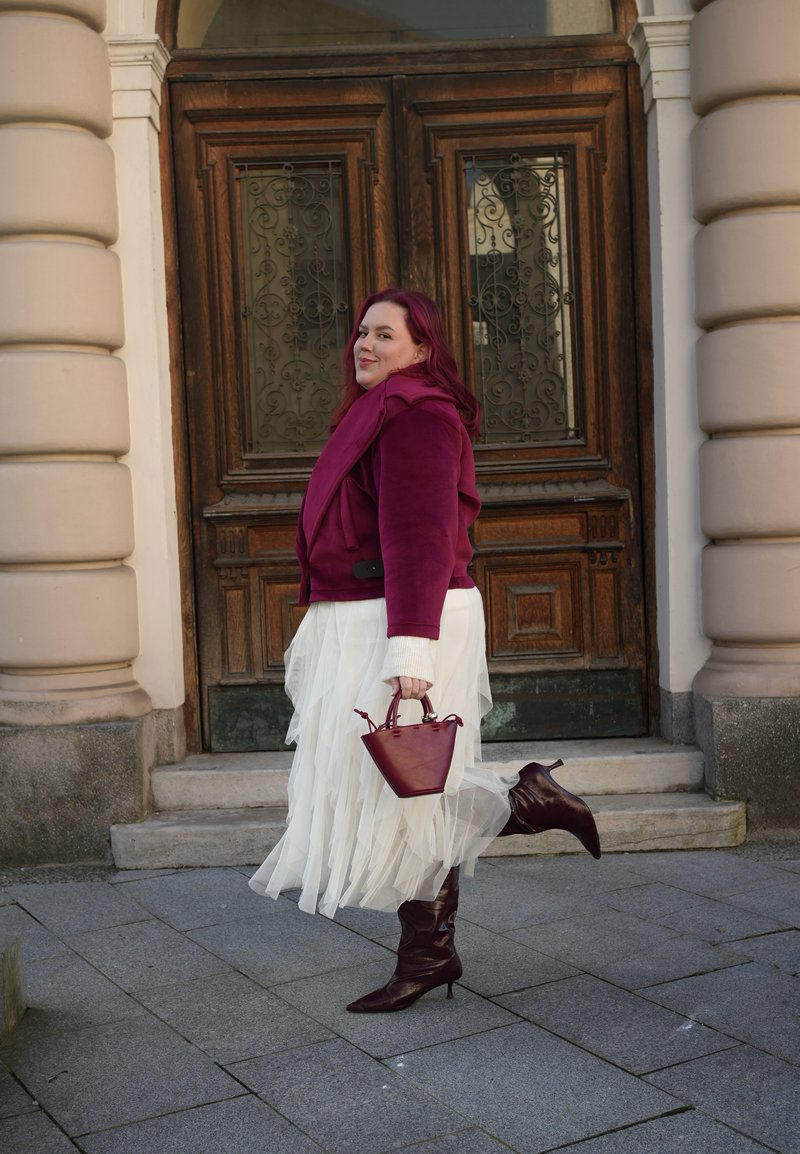 Burgundy suede jacket over a white tulle skirt, paired with burgundy pointed boots and a small matching handbag. Styled outdoors against wooden doors.