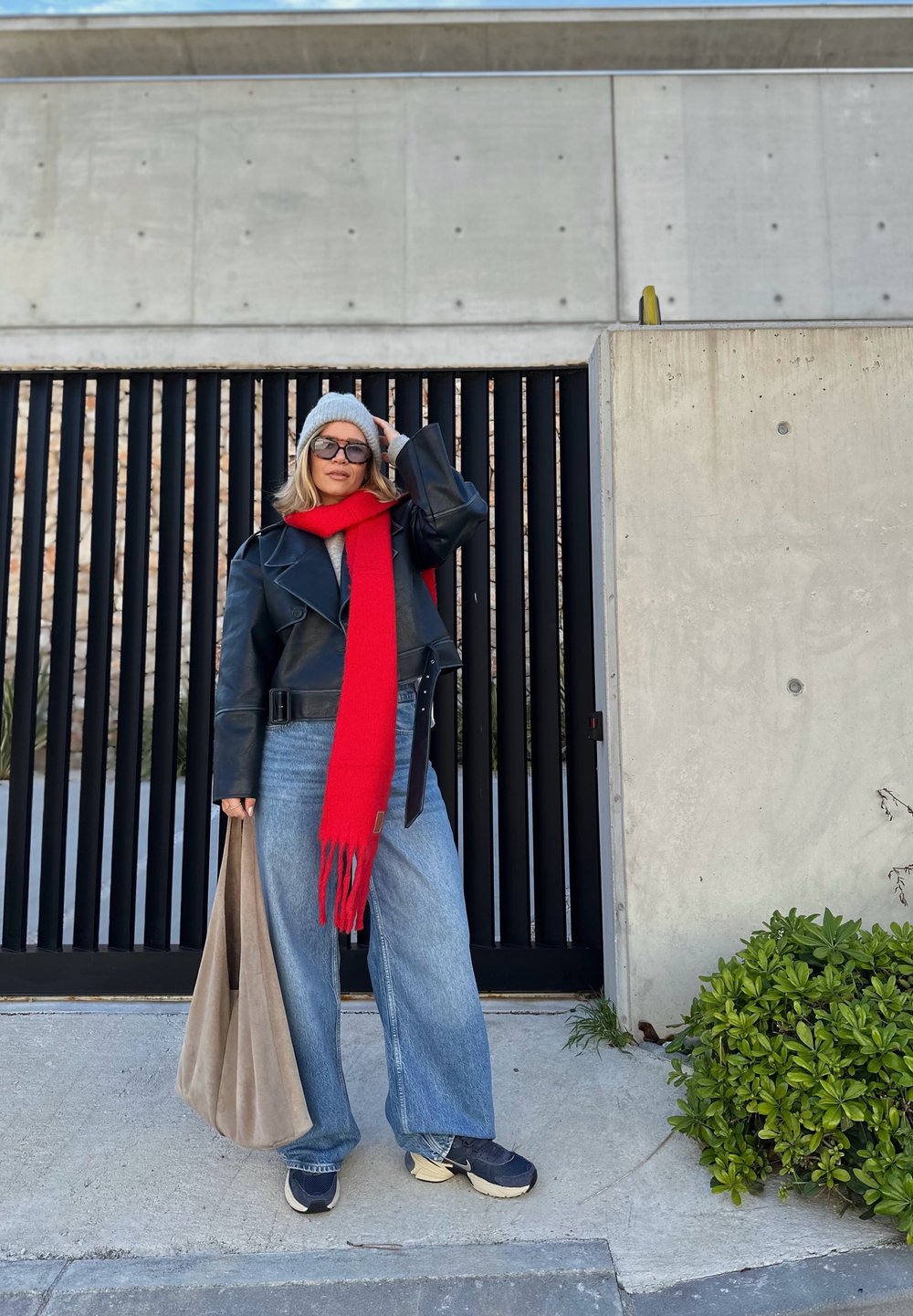 Μαύρο δερμάτινο κοντό μπουφάν, γκρι πουλόβερ, κόκκινο κασκόλ, φαρδιά μπλε τζιν, γκρι σκούφος και αθλητικά παπούτσια; κρατώντας μία μπεζ τσάντα. Σκηνικό από σκυρόδεμα.