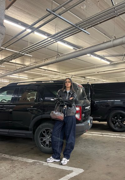 Chaqueta de cuero negra, camiseta gris, pantalones de mezclilla oscuros y holgados, zapatillas deportivas blancas y un pequeño bolso marrón. SUVs negros en un garaje.