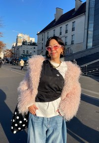 Jeune personne portant une veste rose duveteuse, des lunettes de soleil roses, des hauts noirs et blancs superposés, un jean bleu clair, et tenant un sac noir à pois en plein air.