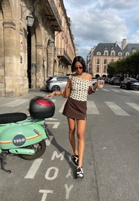 Top branco assimétrico, de um ombro só, com bolinhas pretas; saia mini castanha; sapatos com padrão de pele de vaca; scooter verde ao fundo; ambiente urbano.