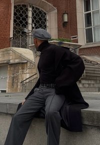 Manteau noir oversize, col roulé noir, pantalon gris ample avec ceinture. Béret gris. En arrière-plan, un mur en brique et une porte ornée.