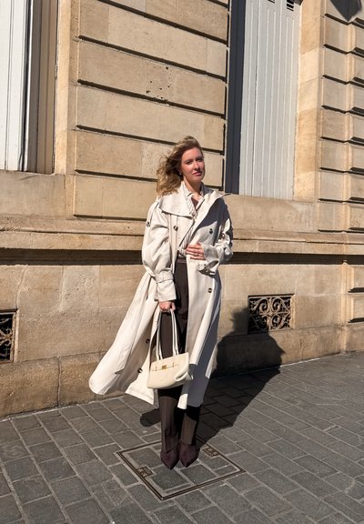 Mujer con cabello largo y rubio, con un impermeable beige fluido, sosteniendo un bolso beige, de pie en una calle empedrada junto a un edificio de piedra.