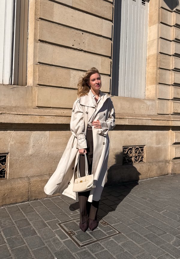 Frau mit langen blonden Haaren in einem fließenden beigen Trenchcoat, die eine beige Handtasche hält und auf einer Kopfsteinpflasterstraße neben einem Steingebäude steht.