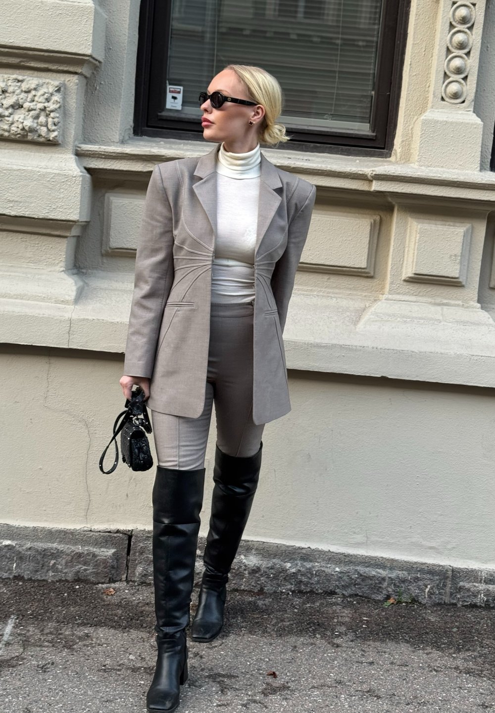 Hall hall kohandatud bleiser tikanddetailidega, valge kõrgekaelusega kampsun, pruunid vormis püksid ja põlveni ulatuvad mustad nahast saapad. Käes must käekott.