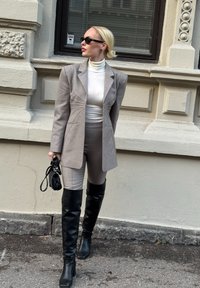 Hall hall kohandatud bleiser tikanddetailidega, valge kõrgekaelusega kampsun, pruunid vormis püksid ja põlveni ulatuvad mustad nahast saapad. Käes must käekott.