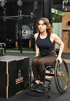 Mujer en silla de ruedas vestida con ropa deportiva preparando para hacer ejercicio junto a una caja pliométrica de madera en un gimnasio.