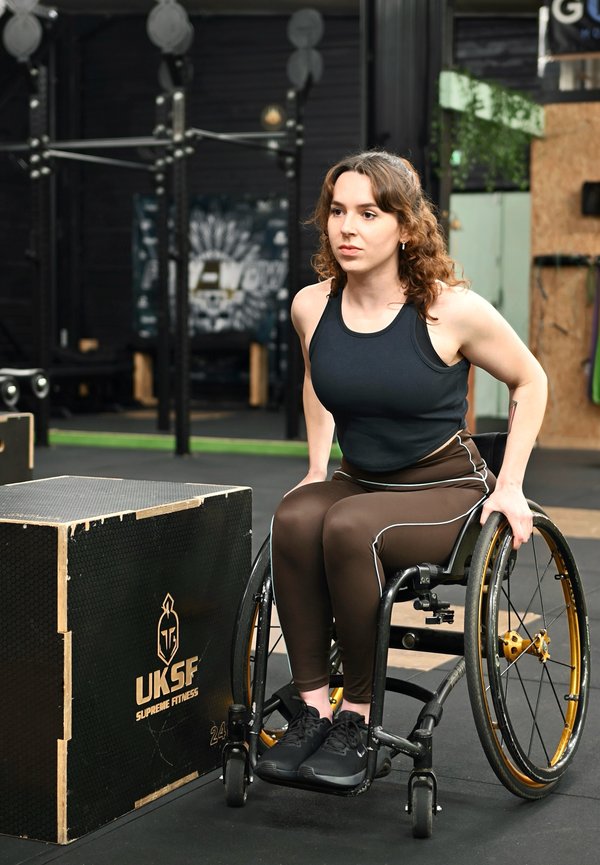 Woman in wheelchair wearing sportswear getting ready to exercise next to a wooden plyometric box in a gym.