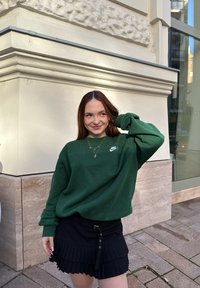 Jovem mulher com cabelo castanho vestindo um sweatshirt verde da Nike e uma saia plissada preta sorri junto a um canto de um edifício, numa calçada pavimentada.