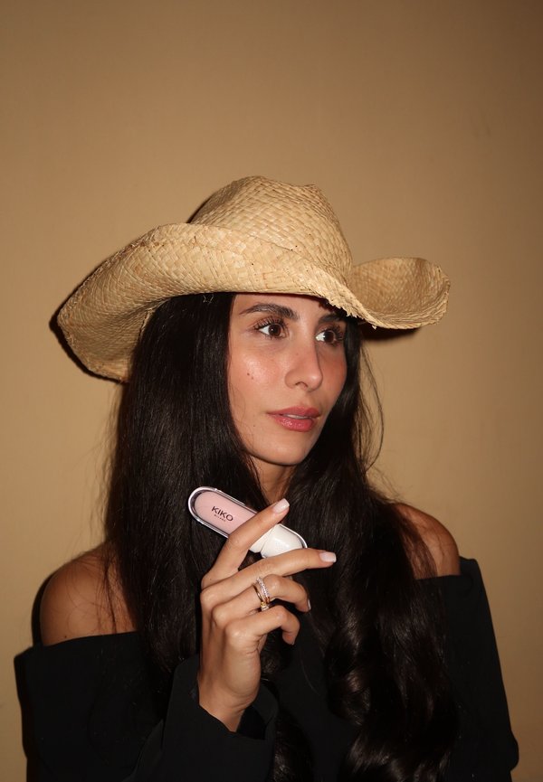 Vrouw met lang, donker haar in een geweven strohoed en een zwarte off-shoulder top, bezig met een roze en witte KIKO Milano cosmetica.