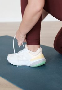 Bílé sportovní boty s žlutým akcentem a zelenou podrážkou, částečně TKD, obuté na noze na modré jóga podložce a burgundských legínách.