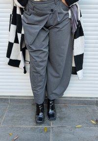 Pantalones grises de corte holgado con pliegue y detalle de botón, combinados con botines negros y brillantes. El fondo muestra una persiana enrollable blanca.