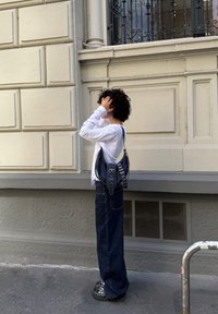 Persoon met krullend haar staat tegen een beige muur, gekleed in een wit langmouwshirt, donkere wijde jeans, zwarte met studs versierde schoenen en een donkere schoudertas.