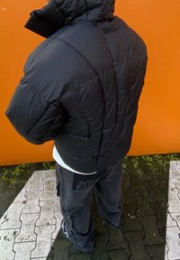 Crna puffer jakna s visokim ovratnikom, prošivenom teksturom i lagano velikim krojem. Nošena preko bijele majice, u kombinaciji s tamnim cargo hlačama.