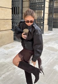 Giacca di pelle nera oversize con spalline, gonna in pizzo trasparente scuro, stivali alti in suede nero, mentre tiene una tazza di caffè.