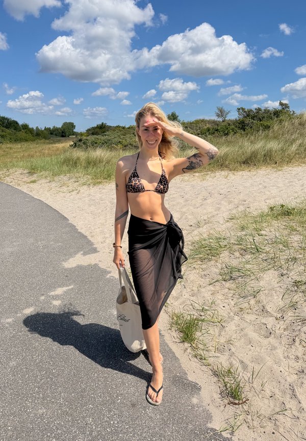 Ung kvinde i leopardprint bikini top og sort sarong, der holder en shopper taske, går ad en solrig strandsti med klitter og blå himmel.