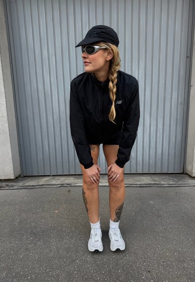 Chaqueta negra ligera de manga larga, usada sobre pantalones cortos negros. Zapatillas deportivas blancas y una gorra negra complementan las gafas de sol espejadas.
