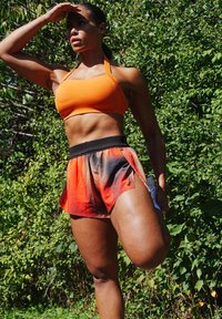 Oranžna halter športna modrček s prilega obliko, v kombinaciji z rdečimi in črnimi prehodnimi tekaškimi kratkimi hlačami z širokim pasom.