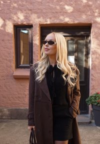 Cappotto lungo in pelle scamosciata marrone, maglione nero a maglia con coulisse, gonna nera, piccoli occhiali da sole ovali, in piedi contro un muro rosa con piante verdi.