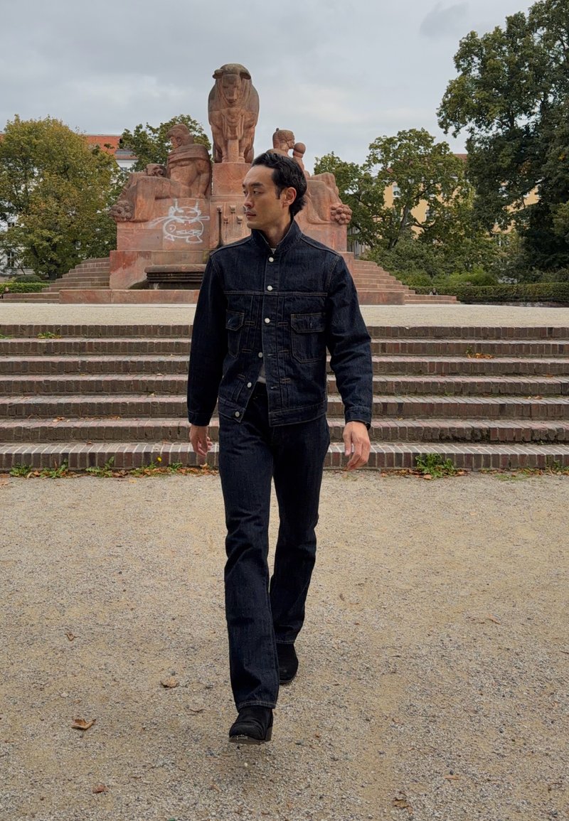 Homme en veste et pantalon en denim foncé marchant sur un chemin de gravier avec des marches en pierre et une grande statue en pierre rouge en arrière-plan entourée d’arbres.