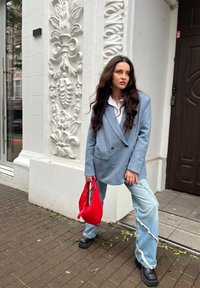 Μοντέλο με ανοιχτό μπλε ριγέ oversized σακάκι, λευκό πουκάμισο, φθαρμένο ανοιχτό μπλε τζιν και ογκώδη μαύρα μποτάκια, κρατώντας μια κόκκινη τσάντα.