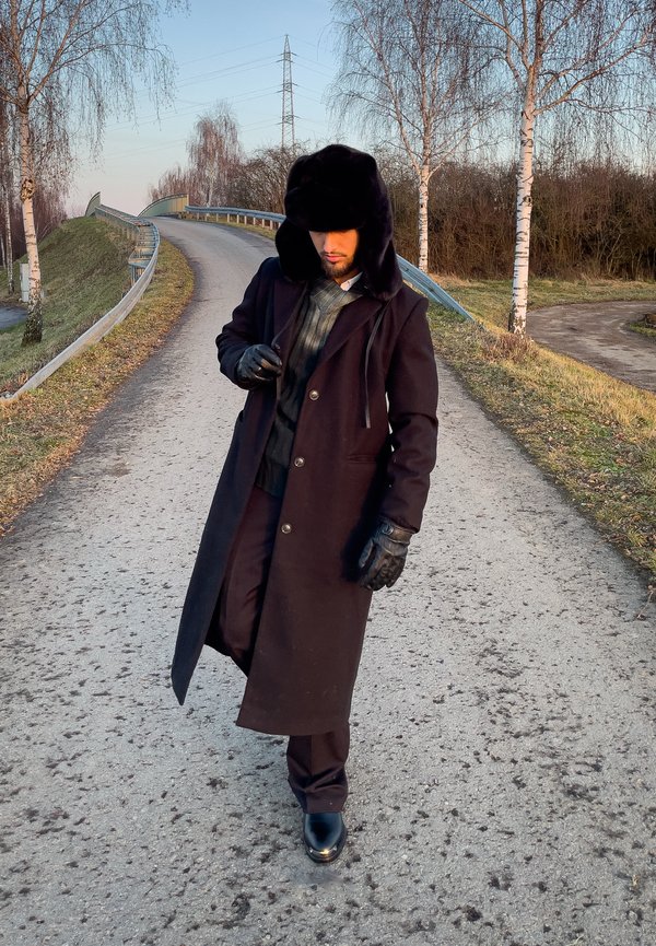 Homme portant un chapeau en fourrure noire, un long manteau foncé, des gants et des bottes, marchant sur une route rurale sinueuse bordée d'arbres dépouillés et de glissières de sécurité.