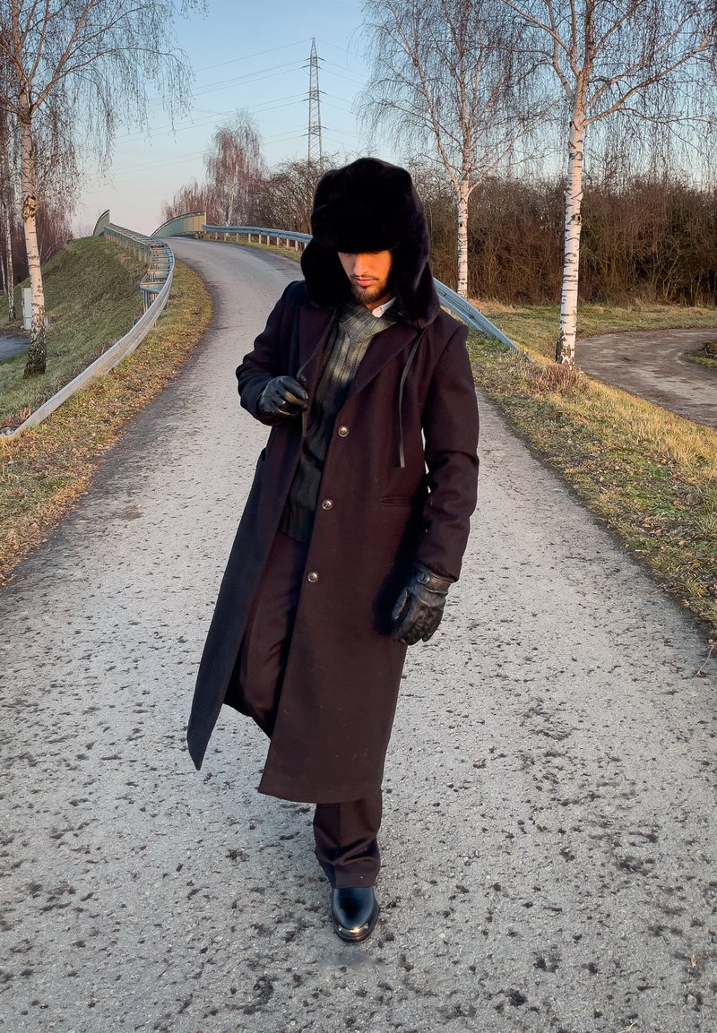 Homme portant un chapeau en fourrure noire, un long manteau foncé, des gants et des bottes, marchant sur une route rurale sinueuse bordée d'arbres dépouillés et de glissières de sécurité.