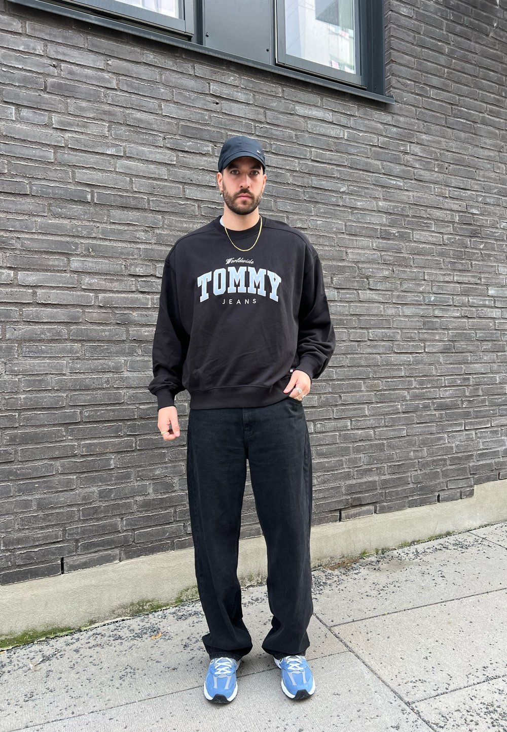 Fekete kapucnis pulcsi "TOMMY JEANS" grafikával világoskék színben, fekete bőszárú nadrággal és kék sportcipővel párosítva, egy téglafal előtt viselve.