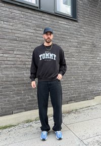 Fekete kapucnis pulcsi "TOMMY JEANS" grafikával világoskék színben, fekete bőszárú nadrággal és kék sportcipővel párosítva, egy téglafal előtt viselve.
