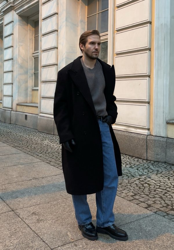 Schwarzer Wollmantel, grauer Rippenpullover, hellblaue Jeans, schwarze Lederhandschuhe und Schuhe. Hintergrund aus Ziegeln und Marmor.