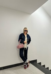 Τζιν τζάκετ, λευκό φουσκωτό μπλουζάκι, σκούρο παντελόνι, ροζ τσάντα χειρός και μπορντώ αθλητικά παπούτσια με λευκές ρίγες, μπροστά σε λευκό τοίχο.