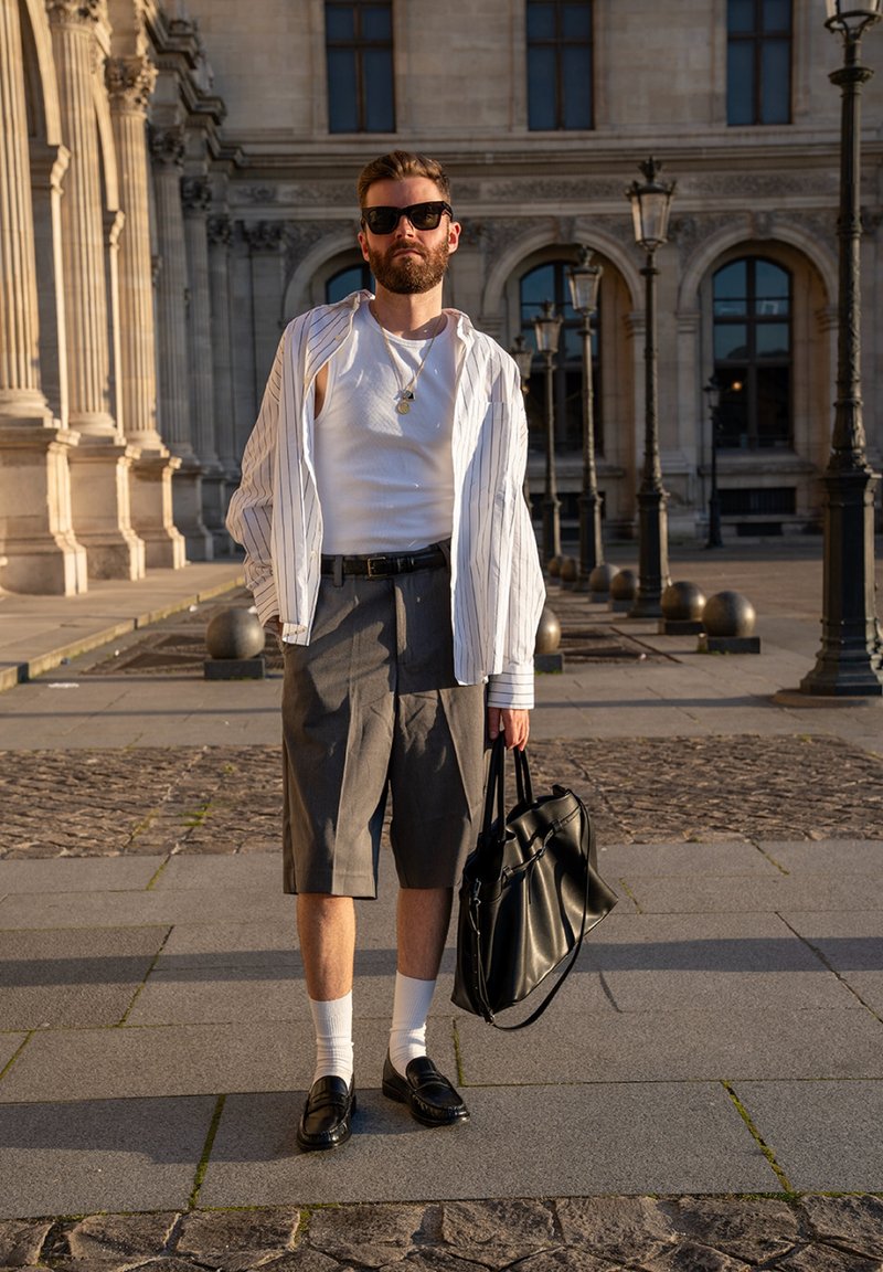 Homme portant des lunettes de soleil, un débardeur blanc, une chemise rayée ouverte, un short gris, des chaussettes blanches et des chaussures noires, tenant un sac noir dans une cour historique.