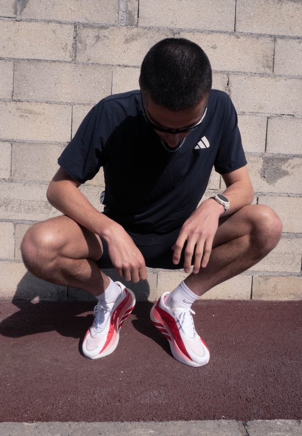 Homme portant une chemise noire, un short noir, des baskets blanches et rouges, et des lunettes de soleil, accroupi contre un mur en briques grises en extérieur.