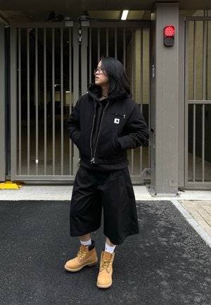 Chaqueta bomber negra con cremallera sobre un sudadera con capucha negra, combinada con pantalones cortos anchos negros y botas de trabajo color beige. De pie cerca de una entrada con reja.