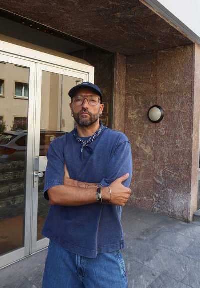Hombre con barba vestido con una camisa azul y pantalones cortos de mezclilla, con los brazos cruzados, cerca de una puerta de cristal y una pared marrón texturizada. Reloj de pulsera casual visible.