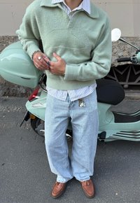 Suéter felpudo verde menta por cima de uma camisa de botão às riscas, combinado com calças de ganga largas em azul claro e sapatos mocassins castanhos, de pé ao lado de uma scooter verde menta.