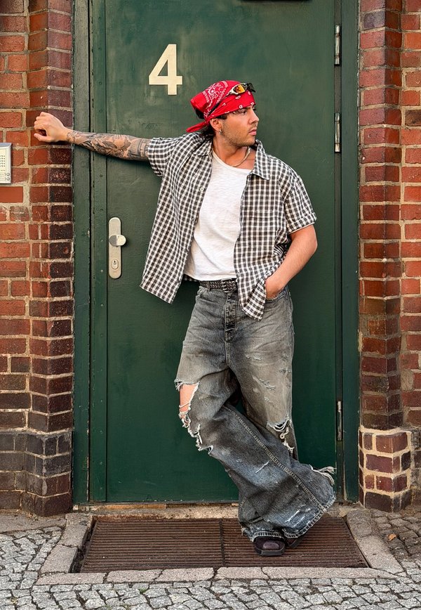 Man in a red bandana and checkered shirt leaning against a green door marked with number 4, wearing ripped wide jeans and sandals.