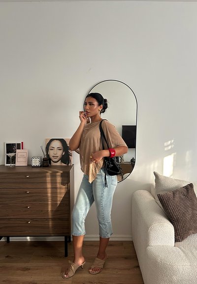 Mujer con una blusa suelta de color marrón claro, jeans cortos de color azul claro, sandalias beige, sosteniendo un pequeño bolso negro. Espejo redondo detrás de ella.