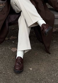 Καφέ δερμάτινα loafers με φούντες, μαύρες καουτσούκ σόλες, συνδυασμένα με λευκές ριγωτές κάλτσες, καθισμένα σε έναν πάγκο με μπεζ φαρδιά παντελόνια.