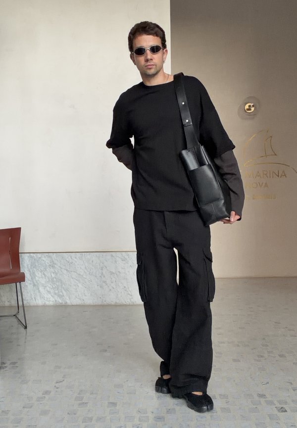 Hombre vestido completamente de negro y con gafas de sol, llevando un bolso negro de hombro, de pie en el interior cerca de una silla y una pared con el cartel "Marina Nova".