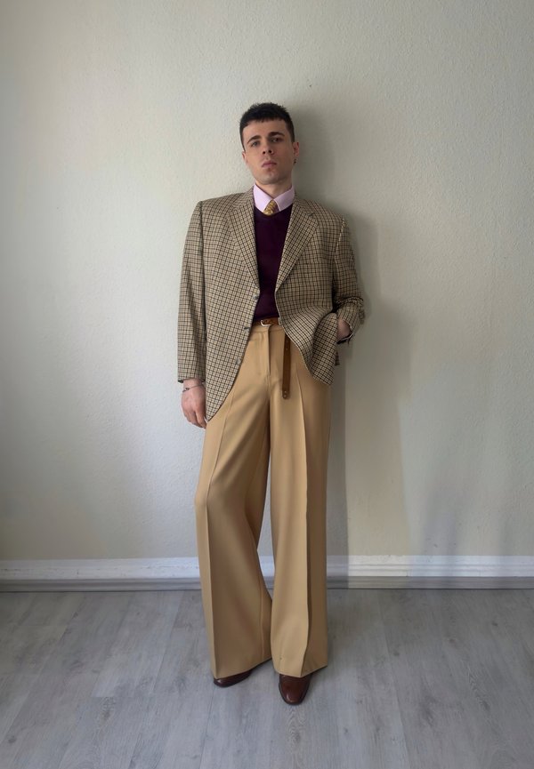 Joven de pie contra una pared beige, vestido con un blazer a cuadros, suéter púrpura, camisa rosa con corbata, pantalones anchos beige y zapatos marrones.