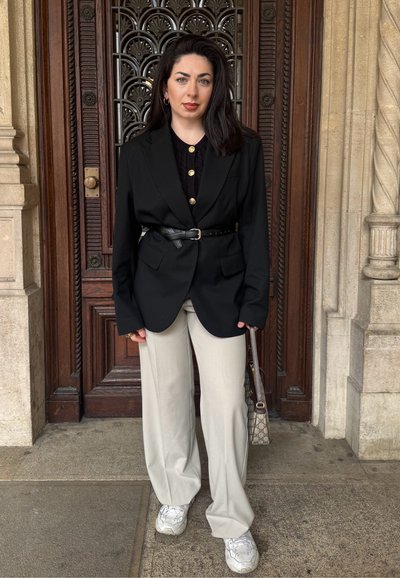 Blazer negro con botones dorados, ceñido a la cintura; combinado con pantalones beiges claros y zapatillas blancas. Fondo de madera texturizada.